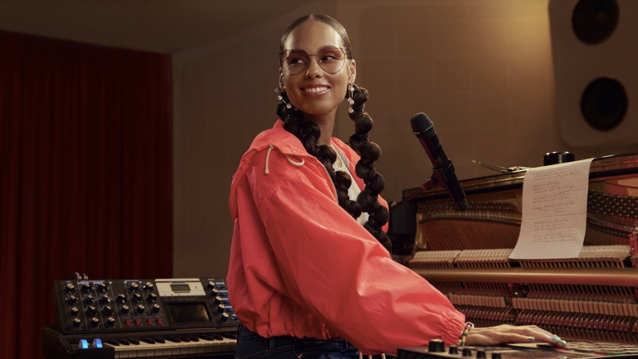 Smiling musician with long braided hair and glasses plays a piano in a recording studio, wearing a bright red jacket, with a microphone and synthesizer nearby and sheet music on the piano
