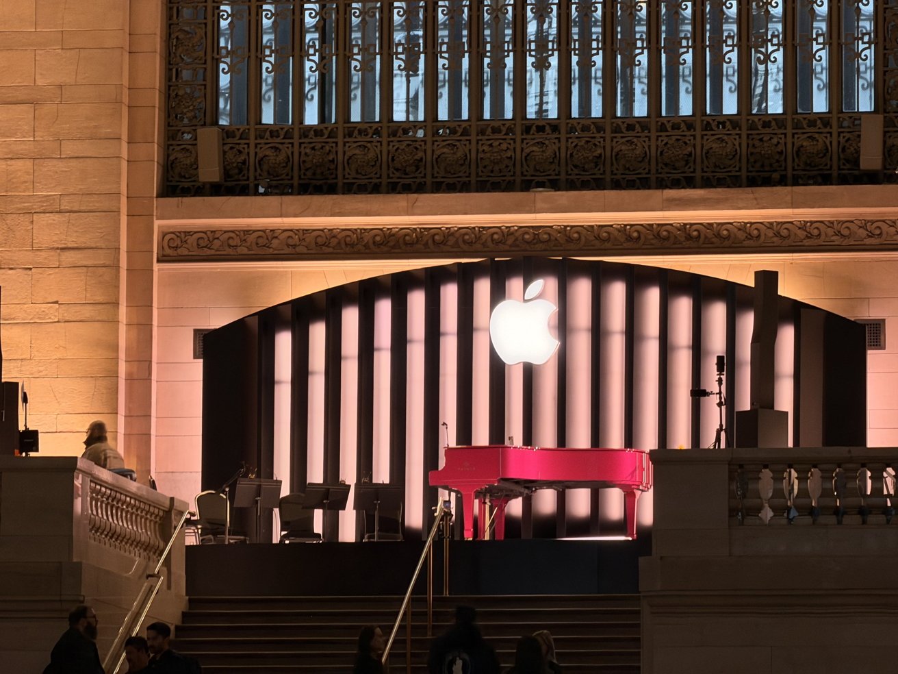 Indoor event setup in a grand hall, featuring a large glowing Apple logo above a lit stage with musical equipment, surrounded by ornate railings and people on nearby staircases