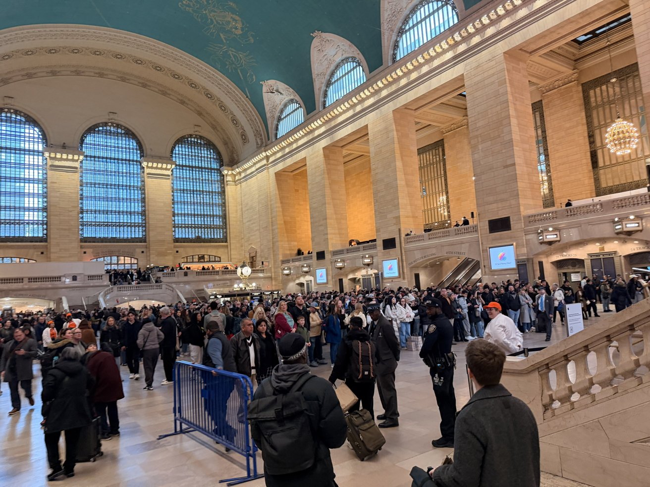 Crowded Grand Central Terminal concourse with commuters, high arched windows, ornate golden lighting, marble walls and staircase, people walking and gathering, blue ceiling partially visible above the bustling scene