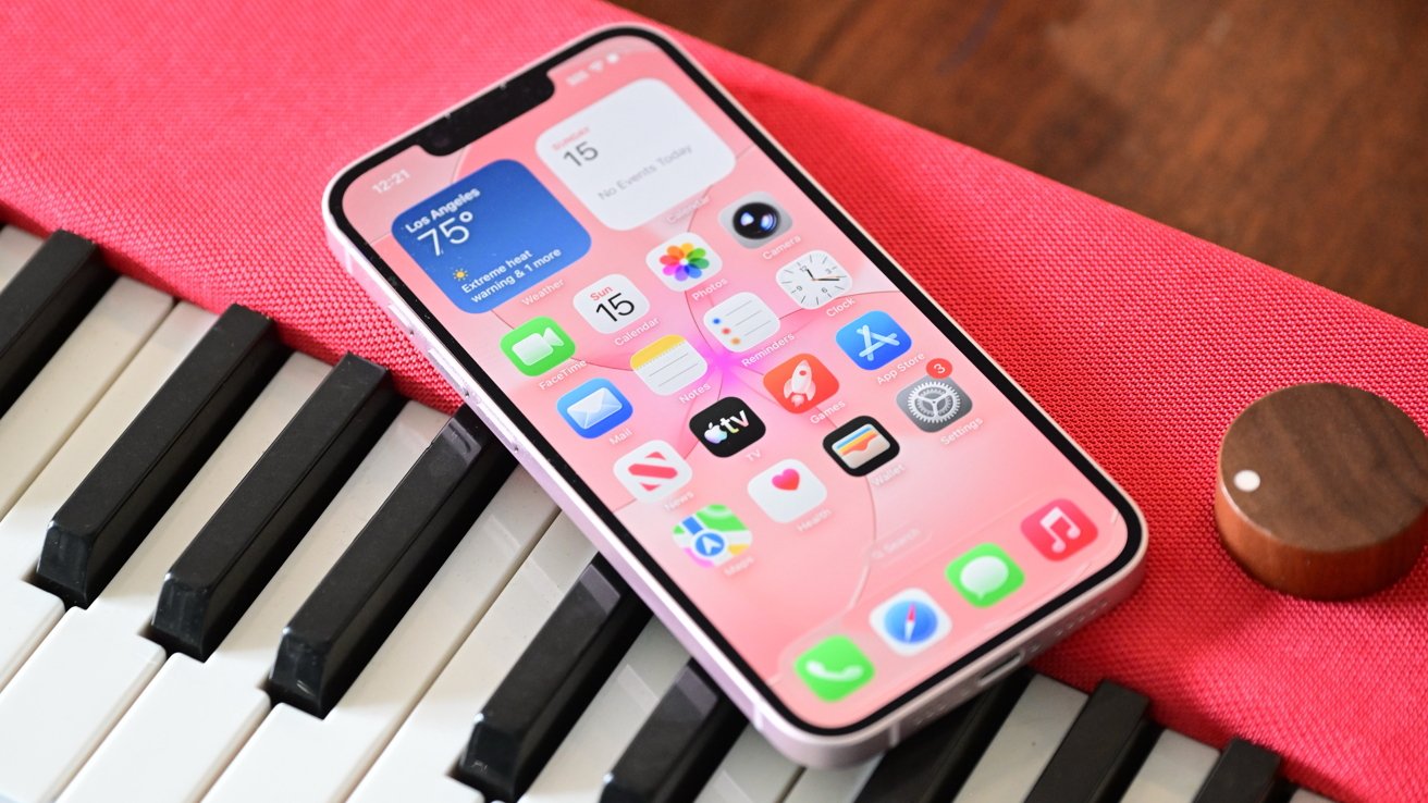 Pink smartphone displaying app icons and weather widget resting diagonally on a piano keyboard, with a bright pink fabric cover and a small wooden knob nearby