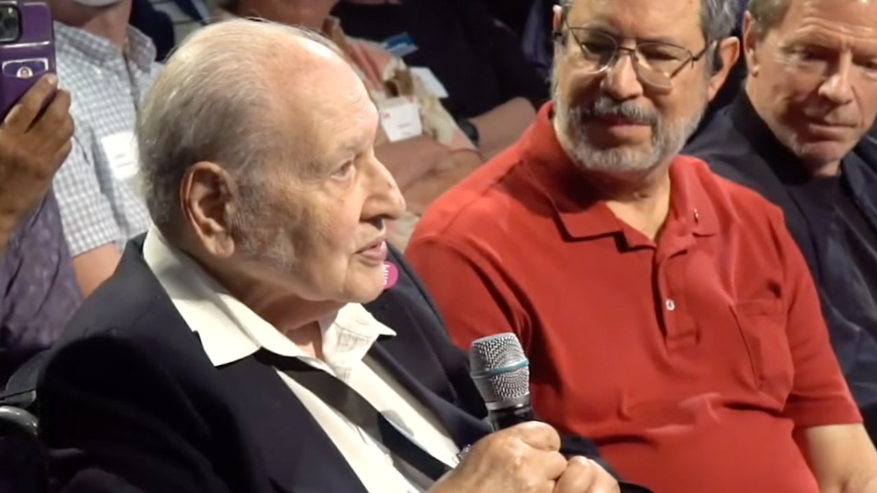 Elderly man in a suit speaks into a microphone at a seated event, surrounded by listening attendees, including a man in a red polo shirt beside him.