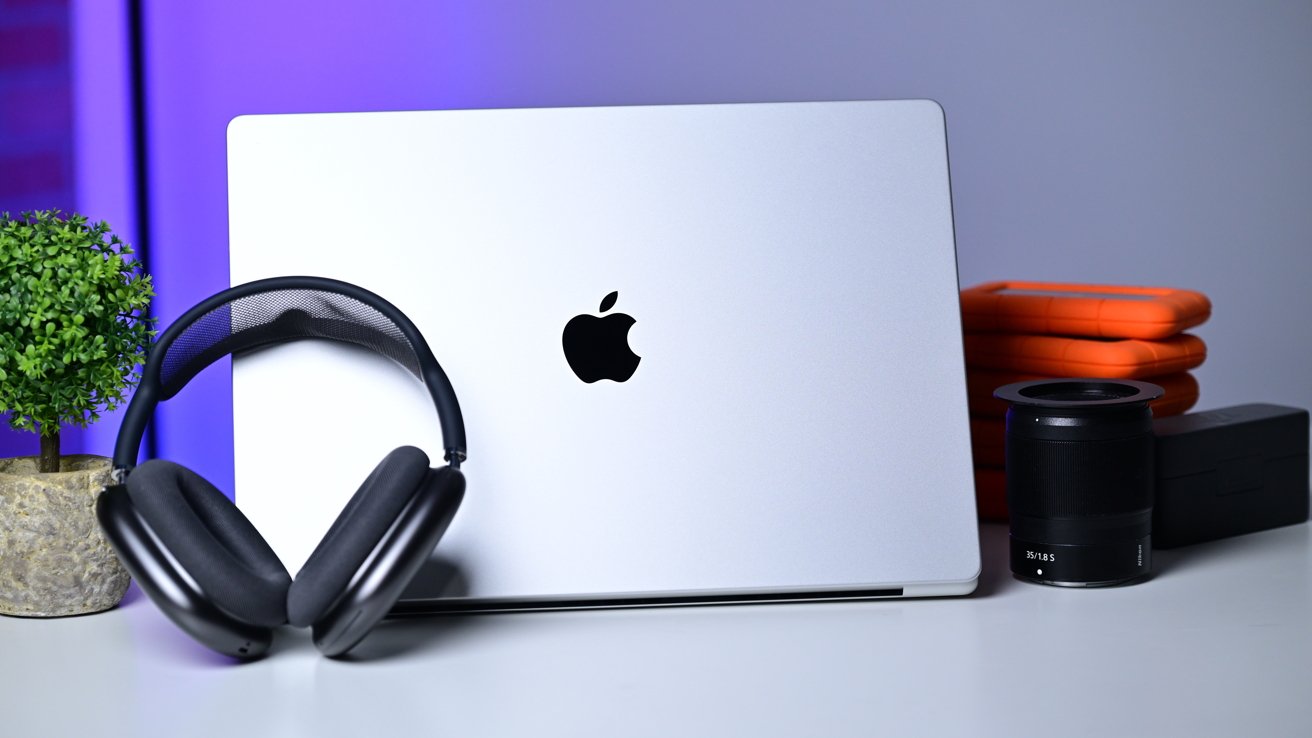 Closed silver MacBook with Apple logo on a desk, black over-ear headphones resting on it, beside a small potted plant, stacked orange hard drives, and a black camera lens.
