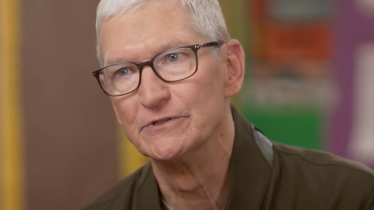 Older man with short gray hair and glasses speaking, shown in close-up indoors with a softly blurred, colorful background.