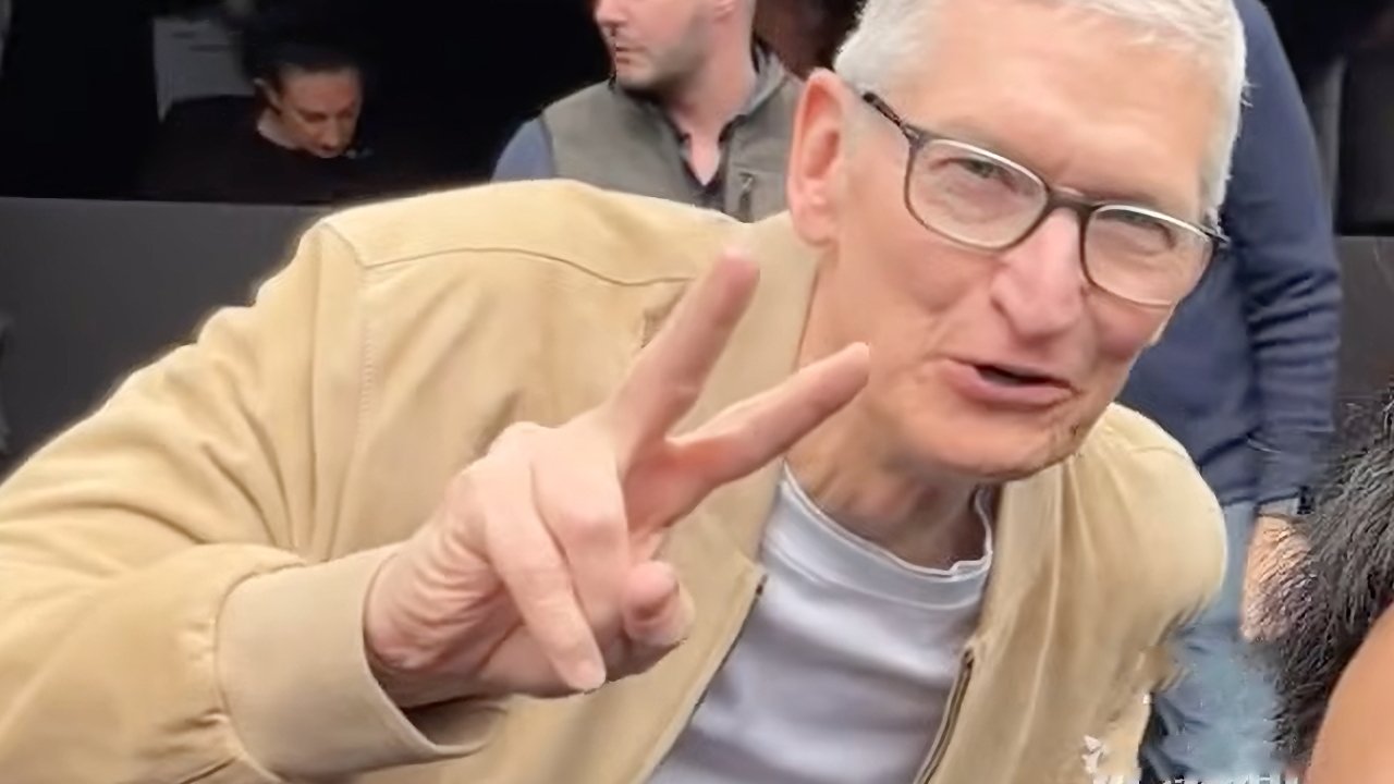 Older man with short gray hair and glasses smiles at the camera, leaning forward and holding up a peace sign with his right hand in a casual, crowded setting