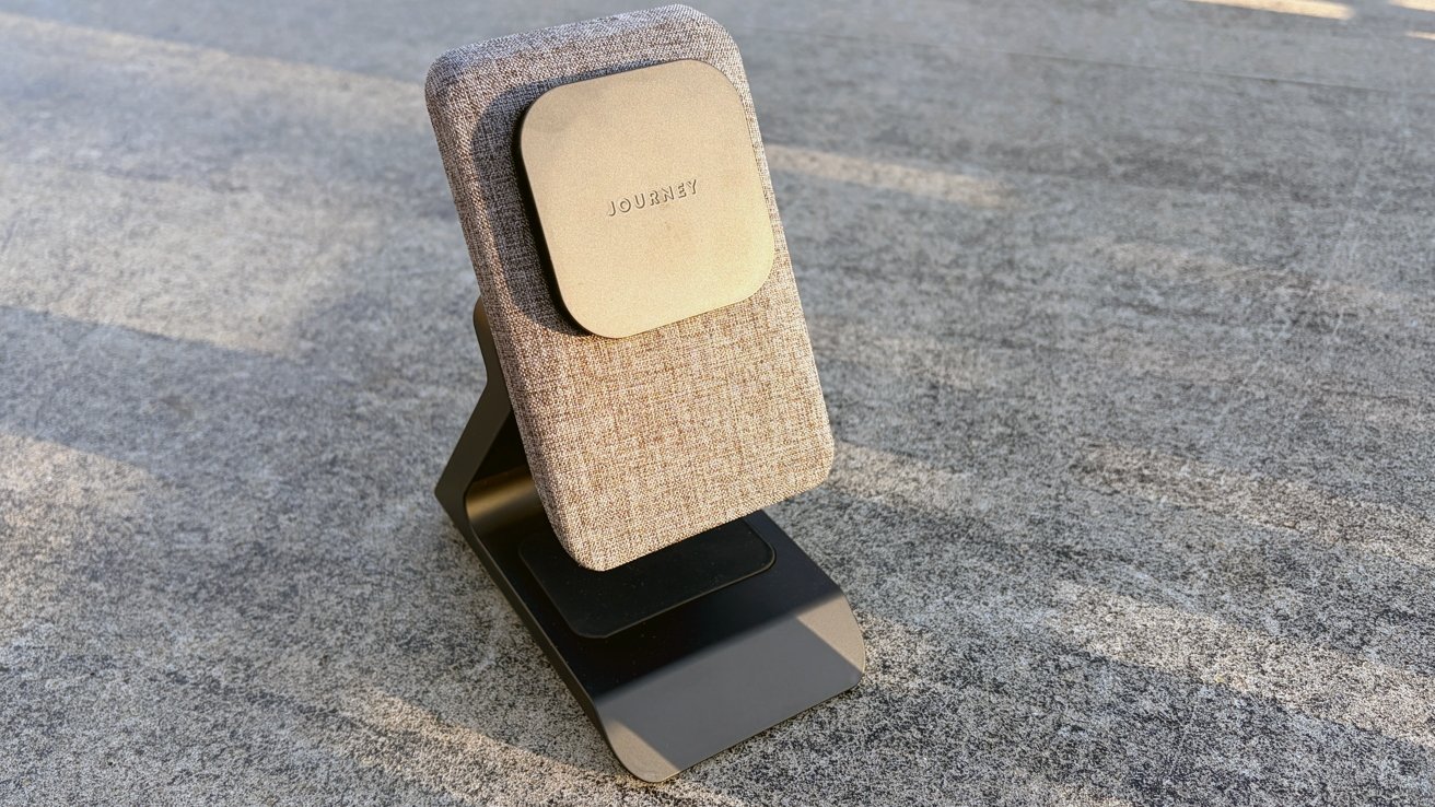 Modern fabric-covered wireless charging stand labeled JOURNEY, standing upright on a flat gray surface in sunlight, with a sleek black metal base and minimalist design