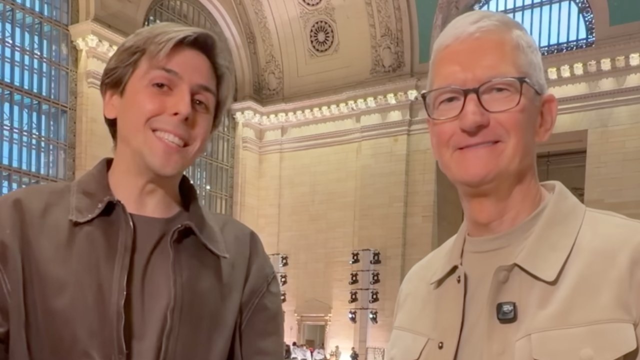 Two smiling men stand close together in a grand hall with tall arched windows, ornate ceiling details, warm lighting, and people gathered in the background.