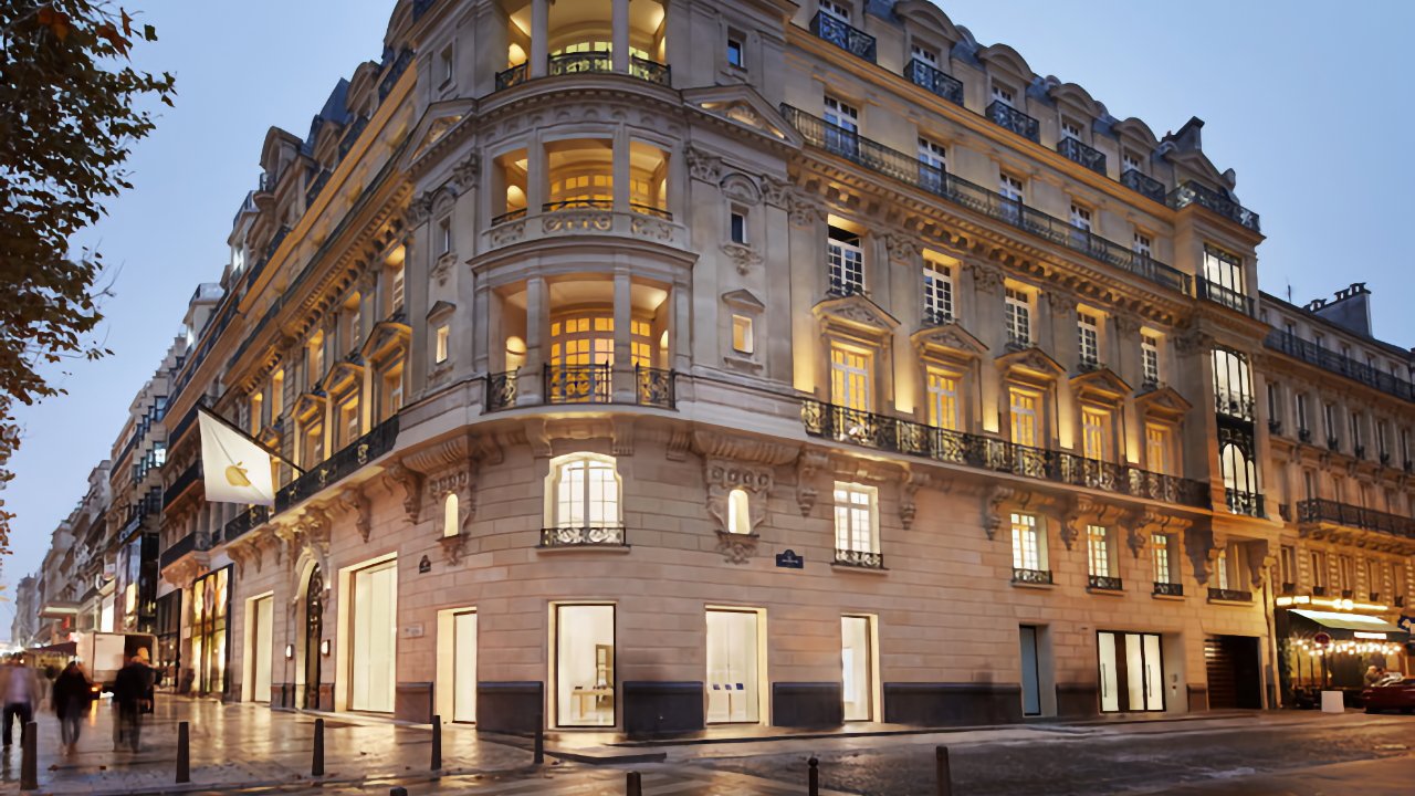 Grand, warmly lit multistory stone building on a city corner at dusk, with ornate balconies, large arched windows, a modern retail storefront at street level, and people walking past.