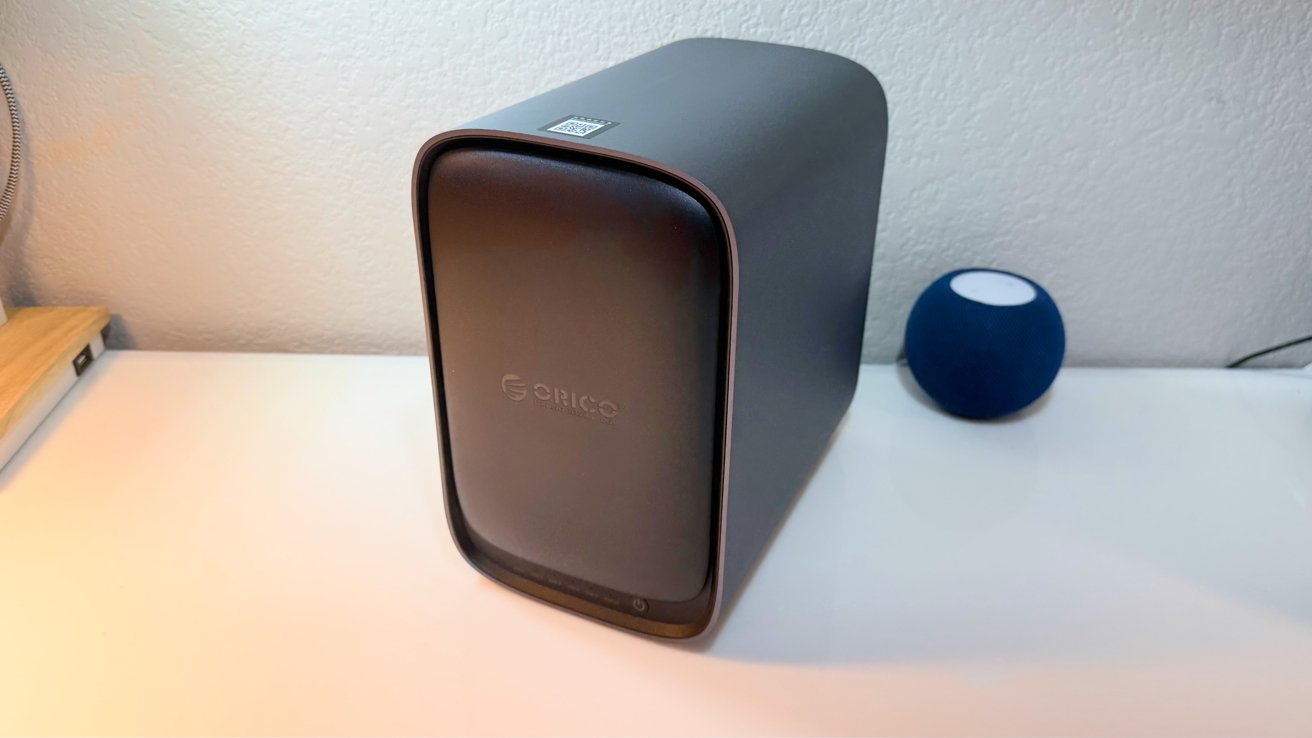 Dark gray Orico external hard drive enclosure on a white desk, with a small blue spherical smart speaker in the background against a light textured wall