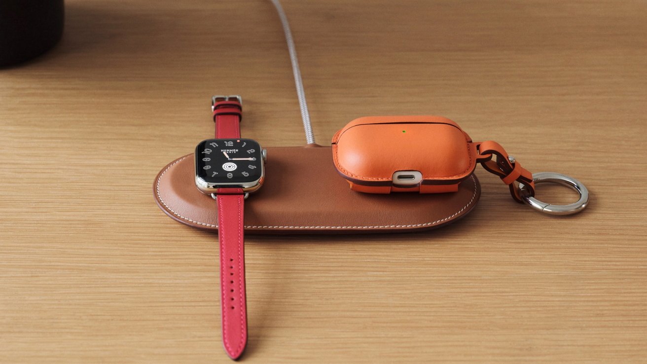Apple Watch with red leather band and orange leather AirPods case on a brown wireless charging pad, resting on a light wooden desk with a metal keyring attached
