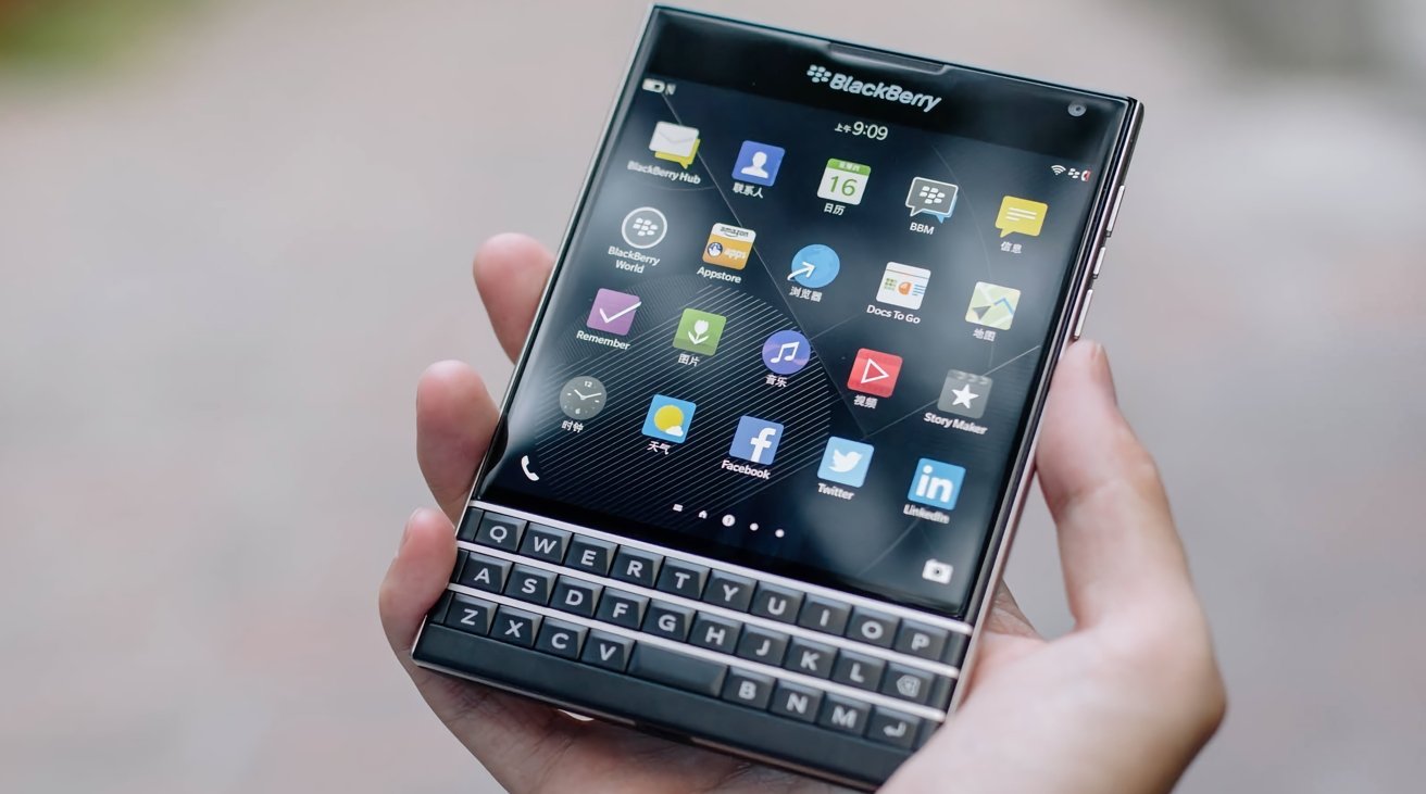 Hand holding a BlackBerry smartphone with square touchscreen, colorful app icons, and a three-row physical QWERTY keyboard, against a softly blurred outdoor background