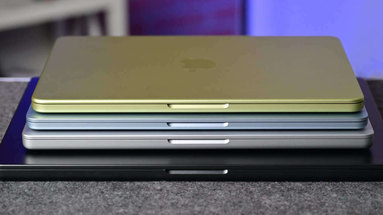 Stack of four closed Apple MacBook laptops in green, blue, silver, and black, aligned neatly on a gray surface with a blurred indoor background