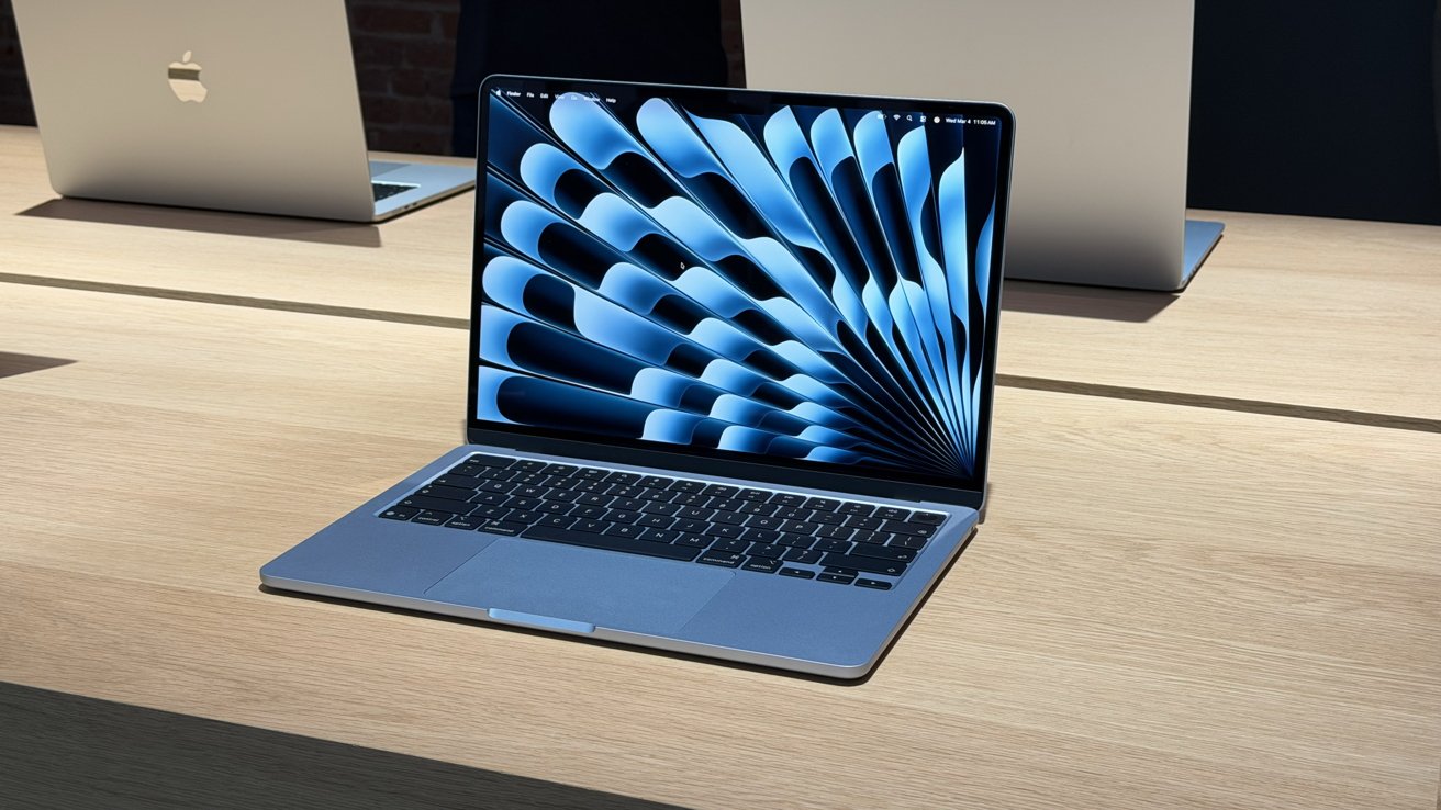 Open MacBook laptop on a light wooden table, displaying a blue abstract radial pattern on its screen, with two larger closed MacBooks positioned in the background.