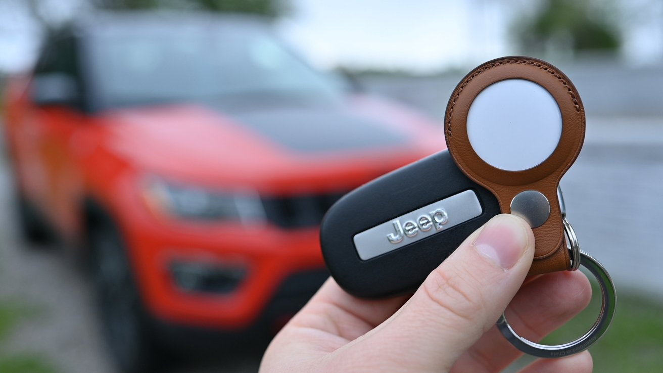 Hand holding a Jeep car key and brown leather tracking tag near a blurred red SUV parked outdoors.