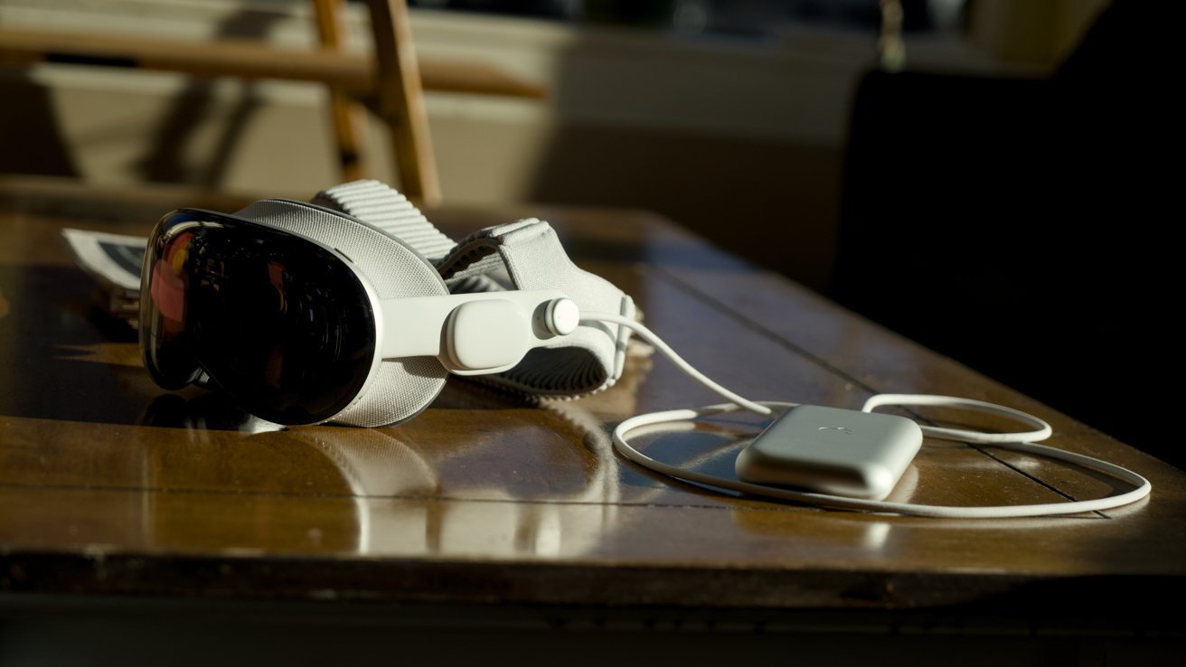 White VR headset with dark reflective visor resting on a wooden table, connected by a cable to a slim rectangular battery pack, in warm indoor sunlight.
