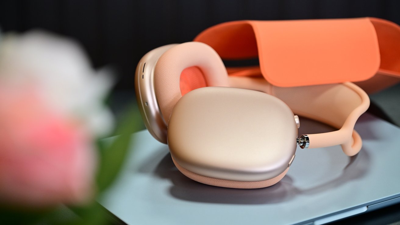 Peach and gold over-ear wireless headphones resting on a reflective surface beside an open matching orange carrying case, with a soft blurred pink flower in the foreground