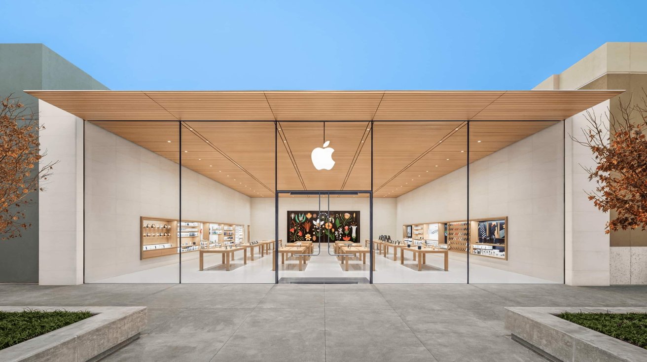 Modern Apple Store exterior with large glass facade, glowing white Apple logo, wooden ceiling, minimalist interior tables displaying products, colorful mural at back, and trees framing the entrance