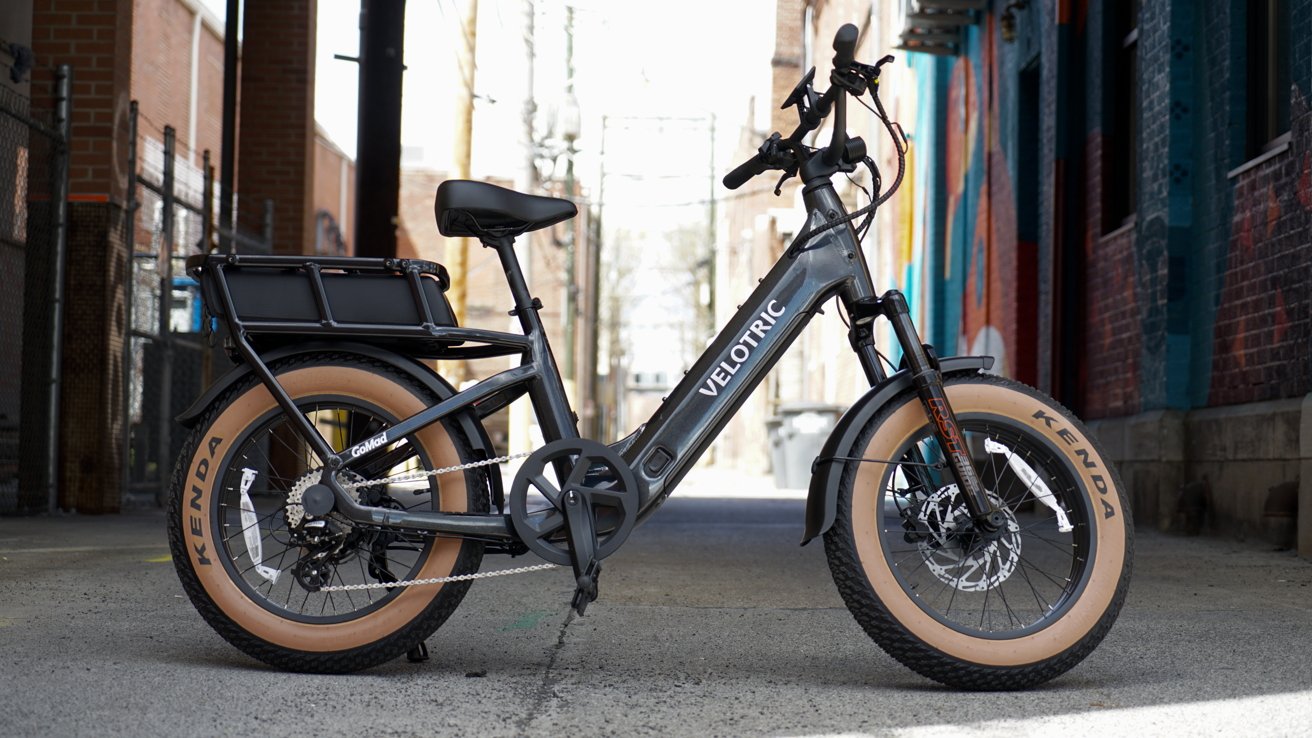 Dark gray Velotric electric bike with tan fat tires parked in a narrow urban alley, showing side profile, rear cargo rack, and front suspension fork