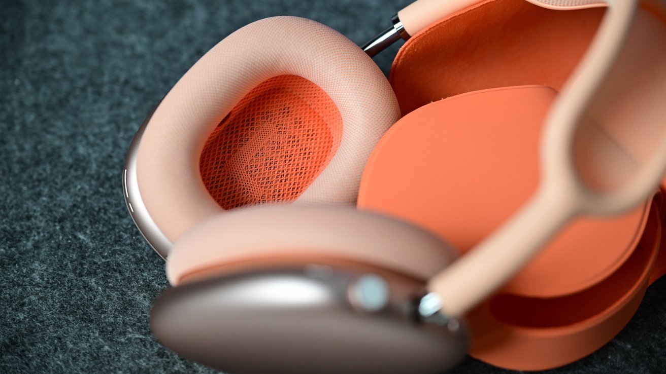 Close-up of peach and orange over-ear headphones resting on a soft dark fabric surface, showing mesh ear cup interior and smooth padded cushions.