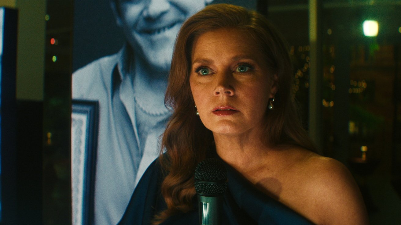 Woman with long wavy hair holds a microphone, looking intensely ahead, dressed in an elegant one-shoulder gown, with a large grayscale portrait of a smiling man behind her.