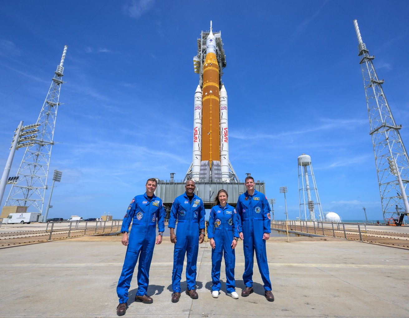 Four astronauts in blue flight suits stand smiling on a launchpad in front of a tall orange and white rocket, under a clear blue sky with support towers around them