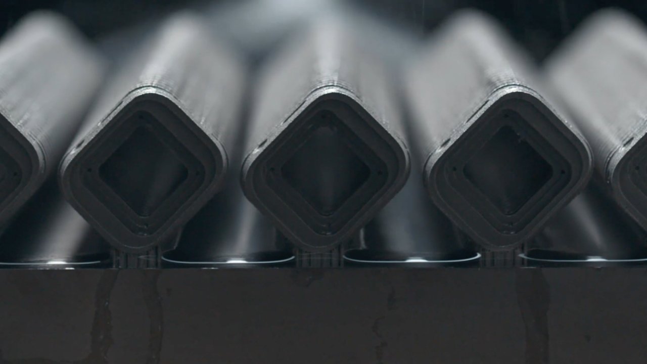 Close-up of stacked hollow metal tubes with square openings arranged in a row, viewed from the front, creating a repeating geometric pattern in dark gray tones