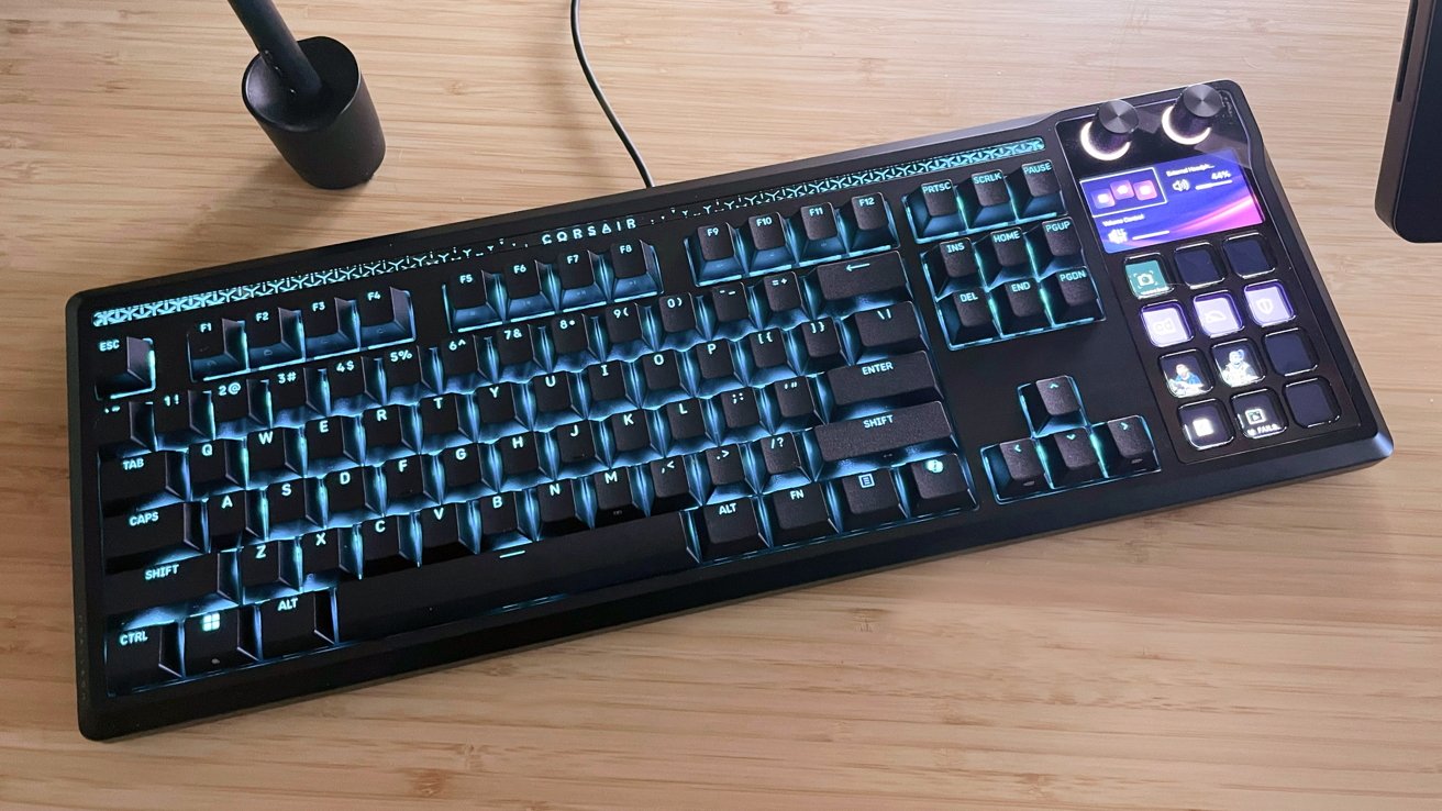 Black Corsair mechanical keyboard with teal backlit keys, integrated right-side macro pad with small screen and icons, two control knobs, on a light wooden desk.
