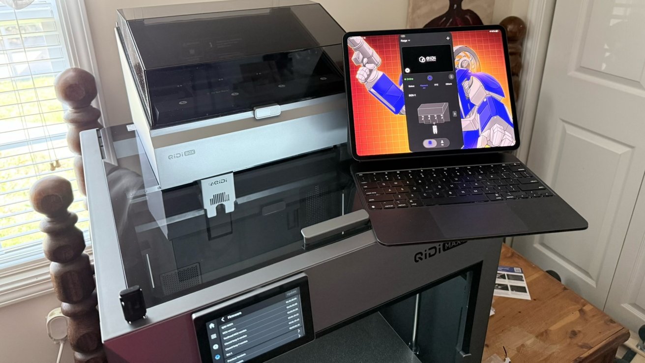 Large enclosed QIDI 3D printer on a wooden table, with a laptop displaying colorful robotic artwork and printer control software, beside a window with blinds and a nearby door