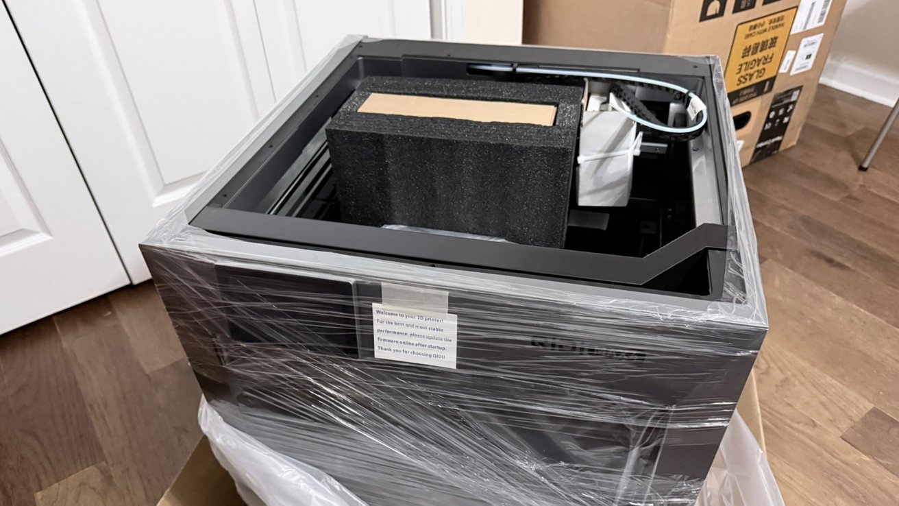 Large 3D printer partially unboxed, wrapped in clear plastic, with a big black foam block and cardboard insert inside the open top, sitting on a wooden floor near boxes