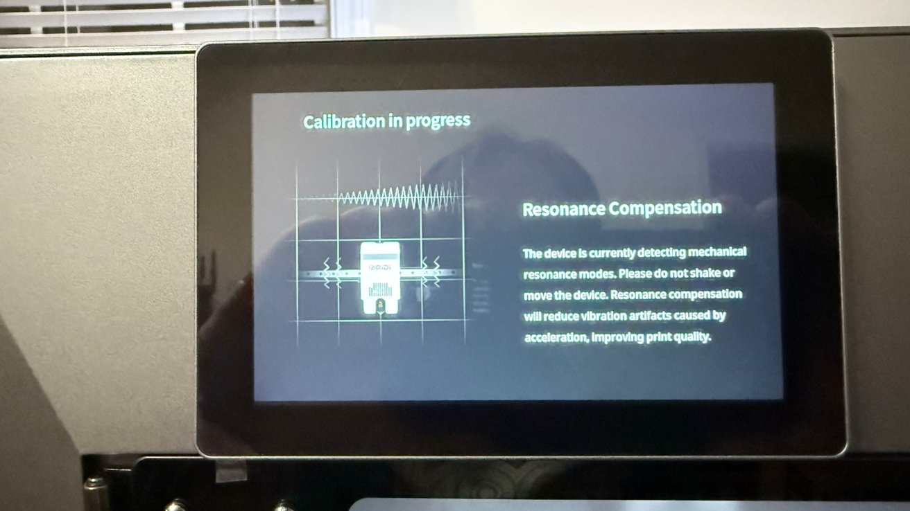 3D printer touchscreen showing calibration in progress for resonance compensation, warning not to move the device while it detects mechanical vibrations to improve print quality