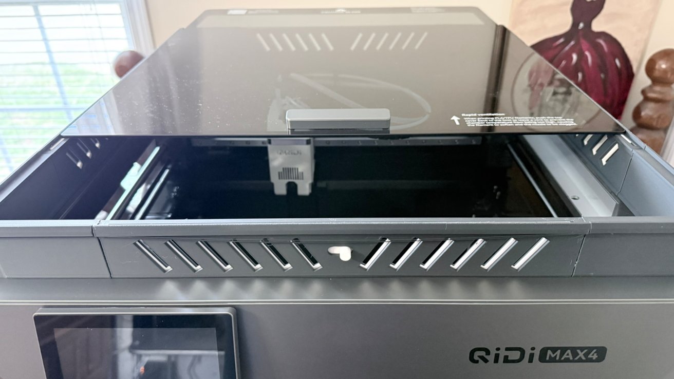 Open-top Qidi Max4 3D printer with lifted dark glass lid, showing interior print area and extruder assembly, positioned indoors near a window with blinds and wall art in background