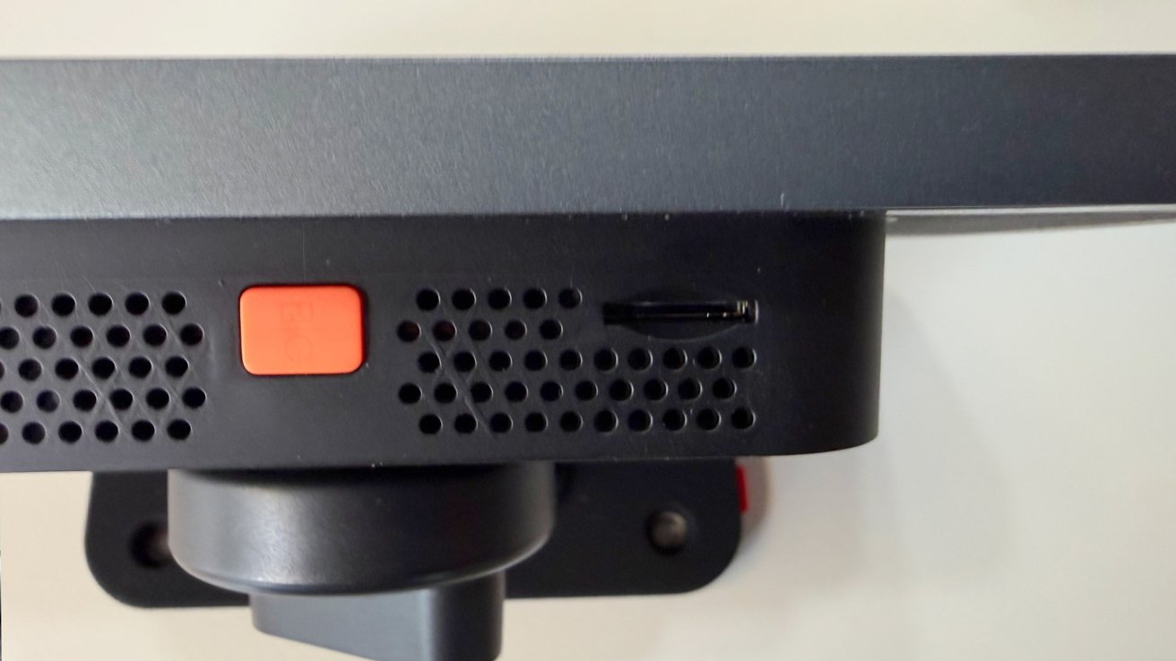 Close-up of a black electronic device with ventilation holes, an orange square button, a small horizontal slot on the right side, and a mounting bracket beneath it against a light background