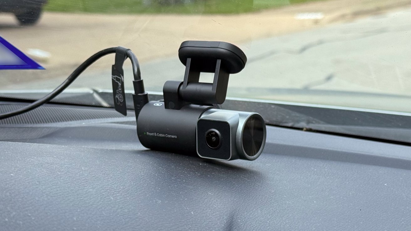 Small black dashcam with cylindrical body and front lens sits on a car dashboard, wired and mounted near the windshield, with a blurry road and grass visible outside.
