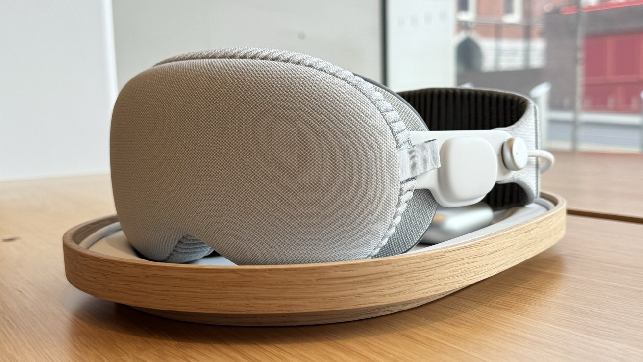 Soft gray padded VR or AR headset resting on a rounded wooden tray atop a light wooden table, with large cushioned front panel and adjustable fabric head strap visible