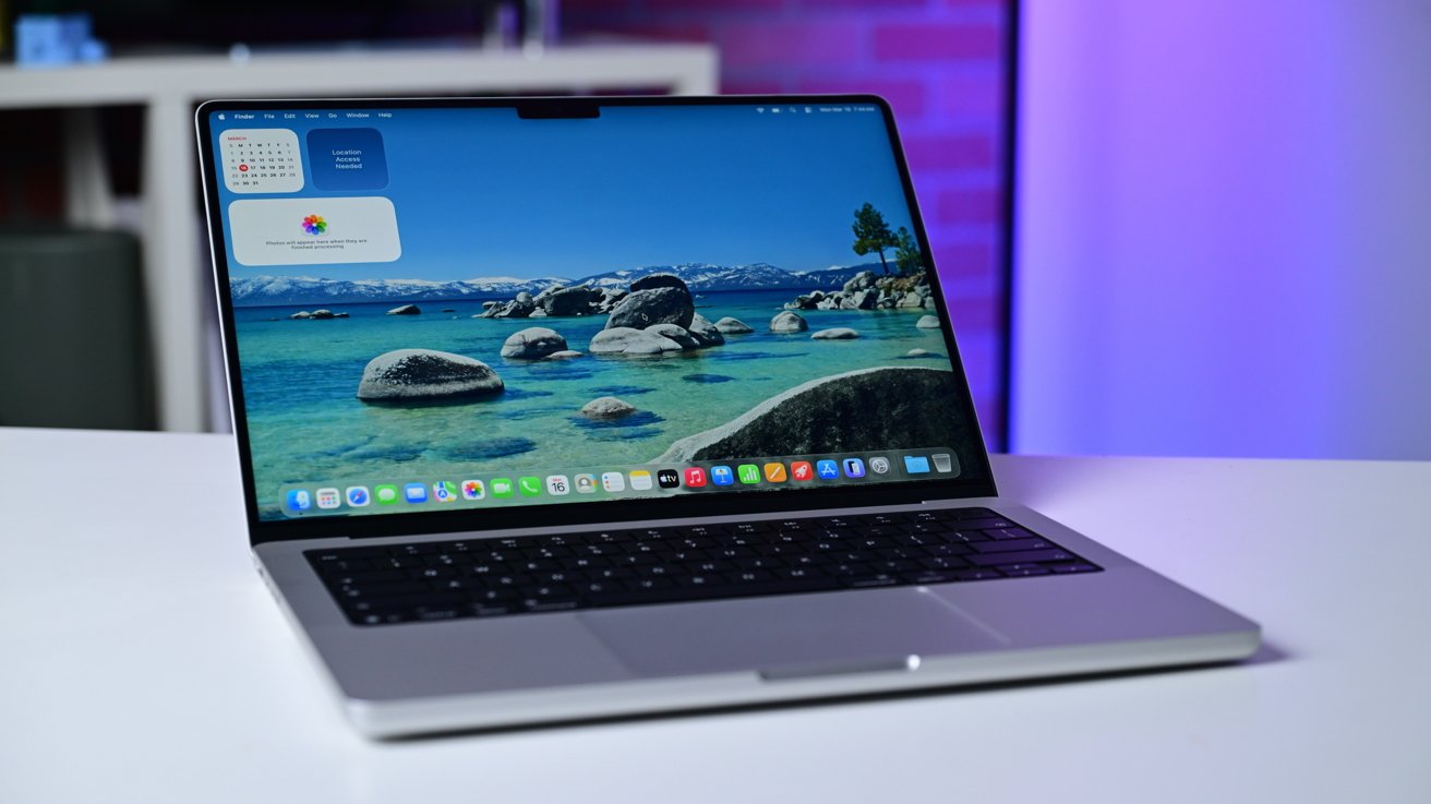 Open MacBook Pro on a white desk, screen showing a vibrant lake and mountains wallpaper with macOS dock icons, calendar widget, and soft purple-blue background lighting