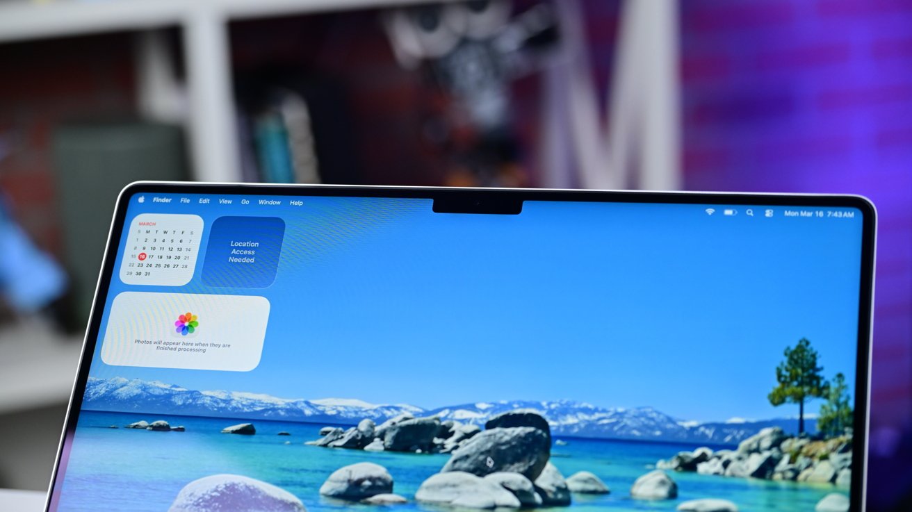 Close-up of a slim laptop screen with a top-center camera notch, showing macOS widgets and a desktop wallpaper of a bright blue lake, rocks, and distant snow-capped mountains.