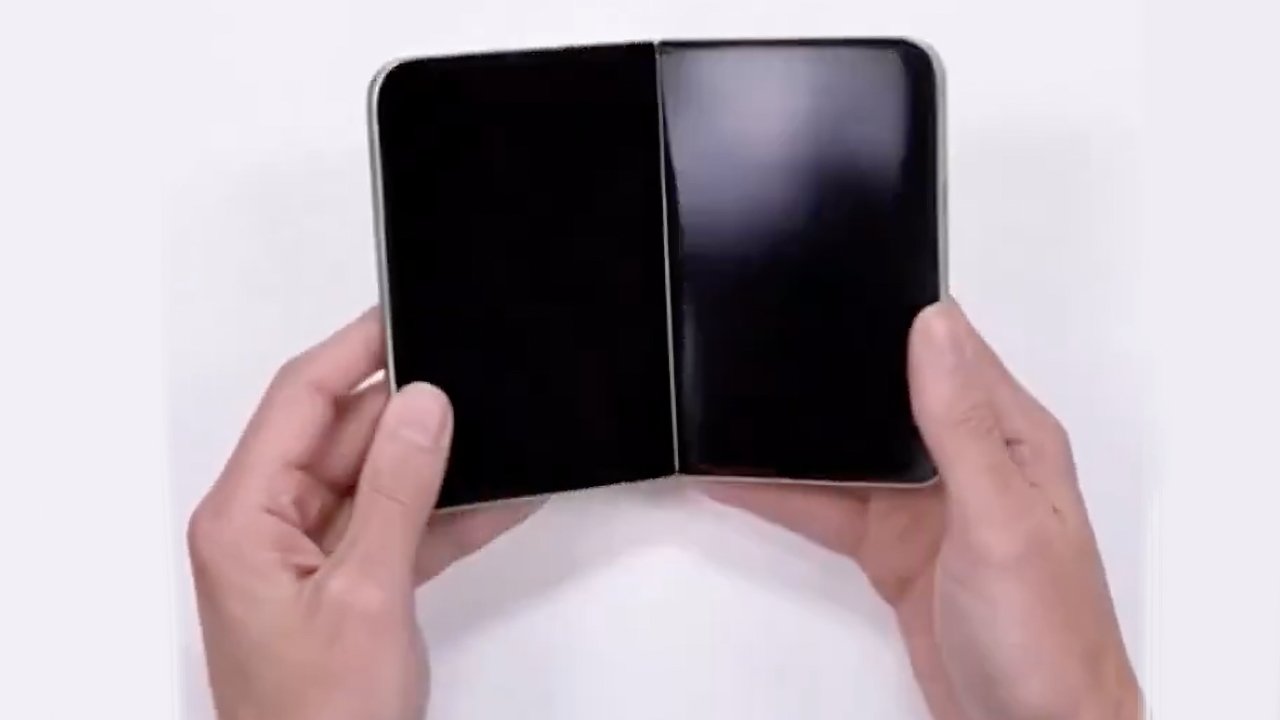 Two hands hold an open foldable smartphone with two blank black screens side by side against a plain light background.