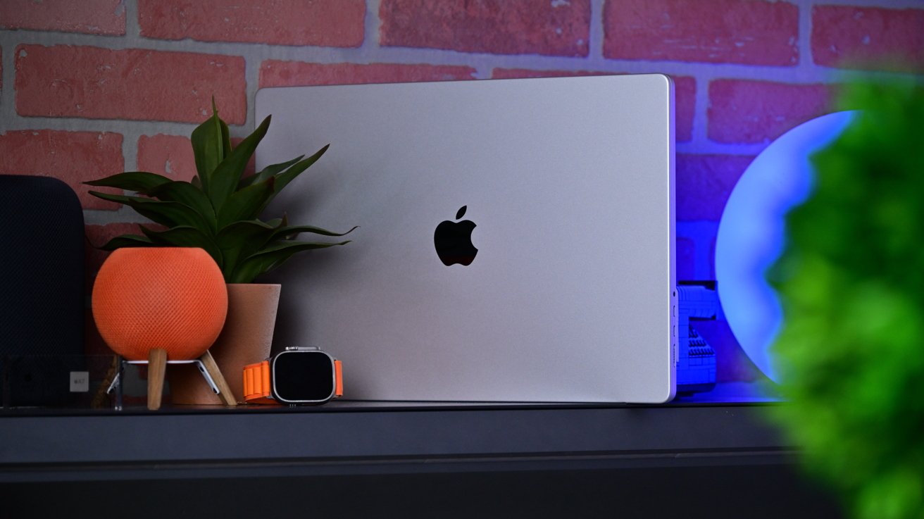 Silver MacBook with Apple logo on a shelf, beside an orange smart speaker, potted plant, Apple Watch with orange band, and glowing blue orb against a brick wall backdrop