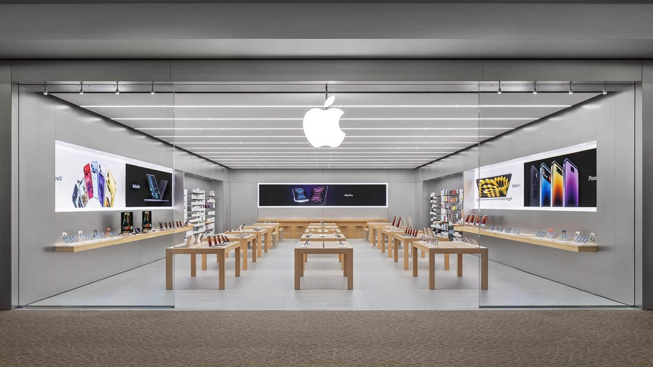 Bright modern Apple retail store with glass front, wooden tables displaying iPhones, iPads, laptops and accessories, illuminated product posters on both walls, and large glowing Apple logo overhead