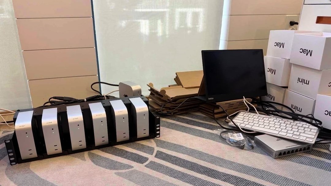 Desk setup with rack-mounted Mac minis, small monitor, white keyboard, Ethernet switch, cables, cardboard trays, and stacked Mac mini boxes in a bright room with window blinds