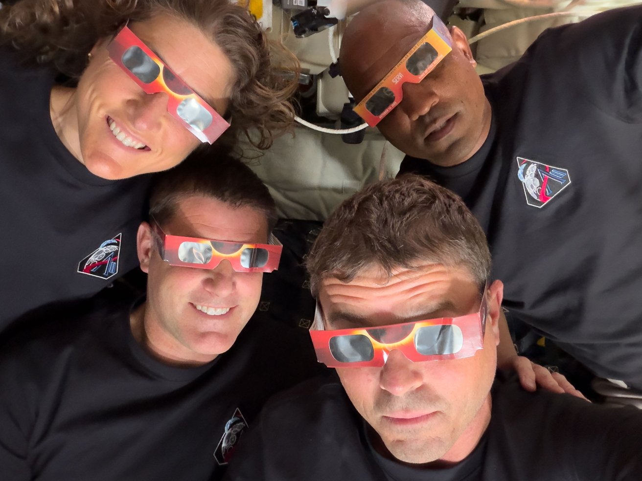 Four astronauts floating close together, smiling and wearing red eclipse glasses, dressed in matching black shirts with mission patches, inside a spacecraft cabin filled with equipment.