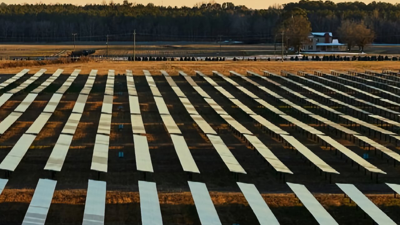 Rows of reflective solar panel arrays stretch across a flat rural field at sunset, with a distant farmhouse, trees, and power lines along the horizon.