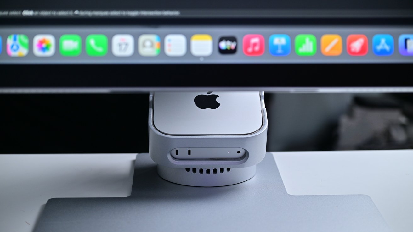 Silver Mac mini computer docked beneath an Apple monitor displaying colorful macOS app icons, all placed on a sleek gray desktop stand in a modern workspace