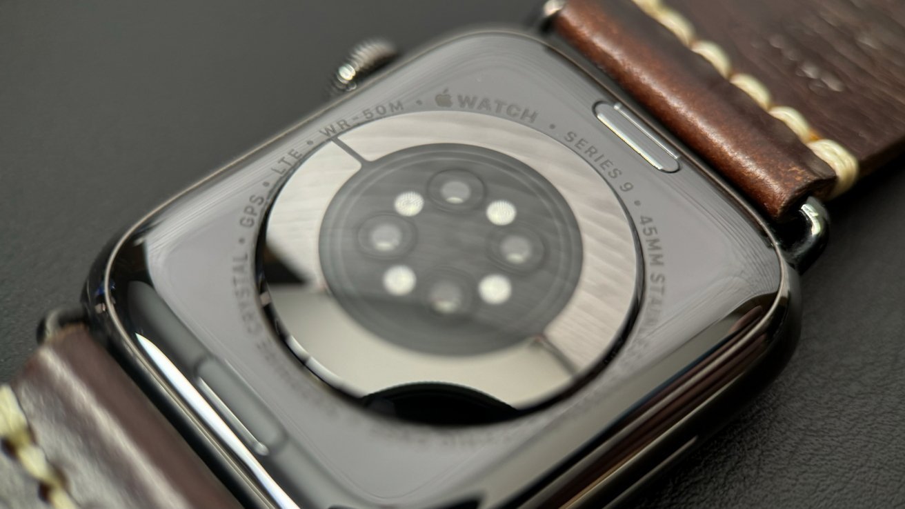 Close-up of an Apple Watch Series 9 back, showing circular heart-rate sensor cluster and engraved text, attached to a brown leather band with visible stitching on a dark surface