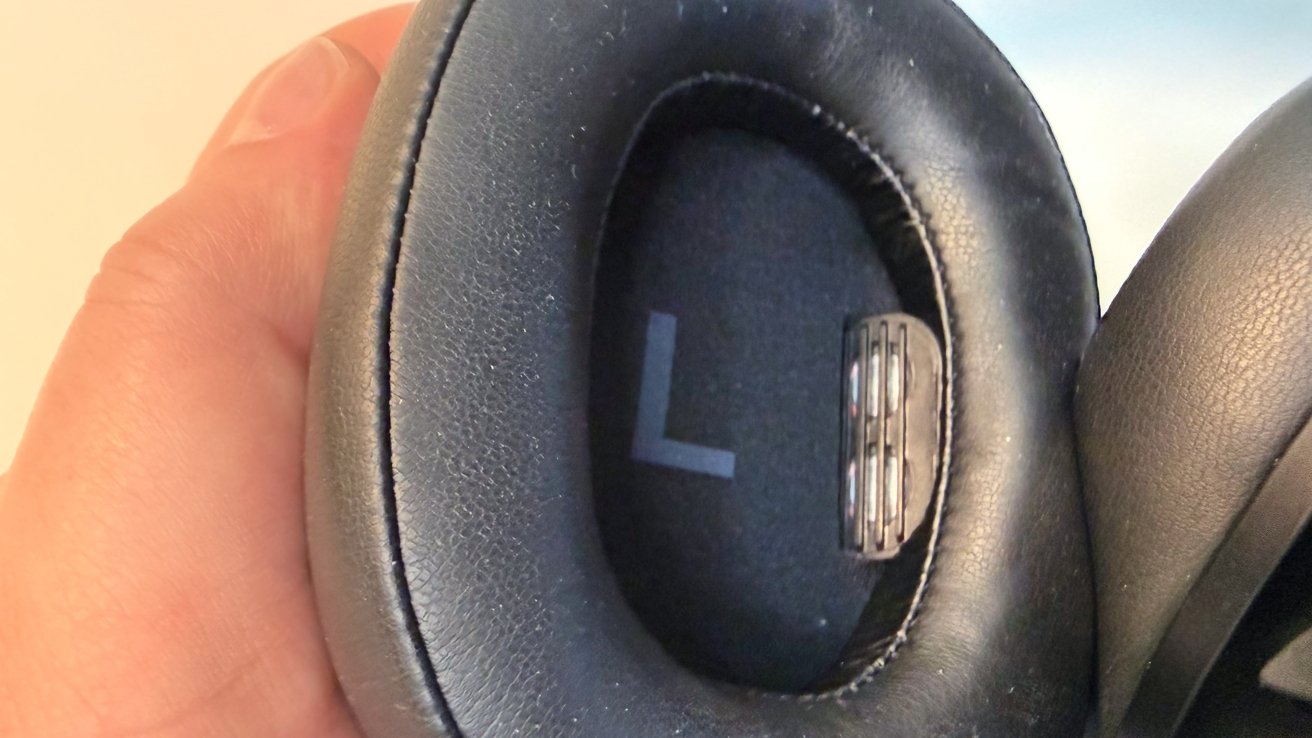 Close-up of a hand holding a black over-ear headphone cushion, showing the large letter L inside the ear cup and a small vented plastic component on the side.