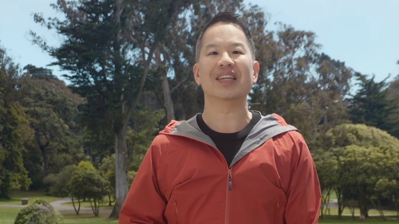 Man in an orange jacket smiles while standing in a sunny park, surrounded by tall trees, green grass, and a clear blue sky in the background