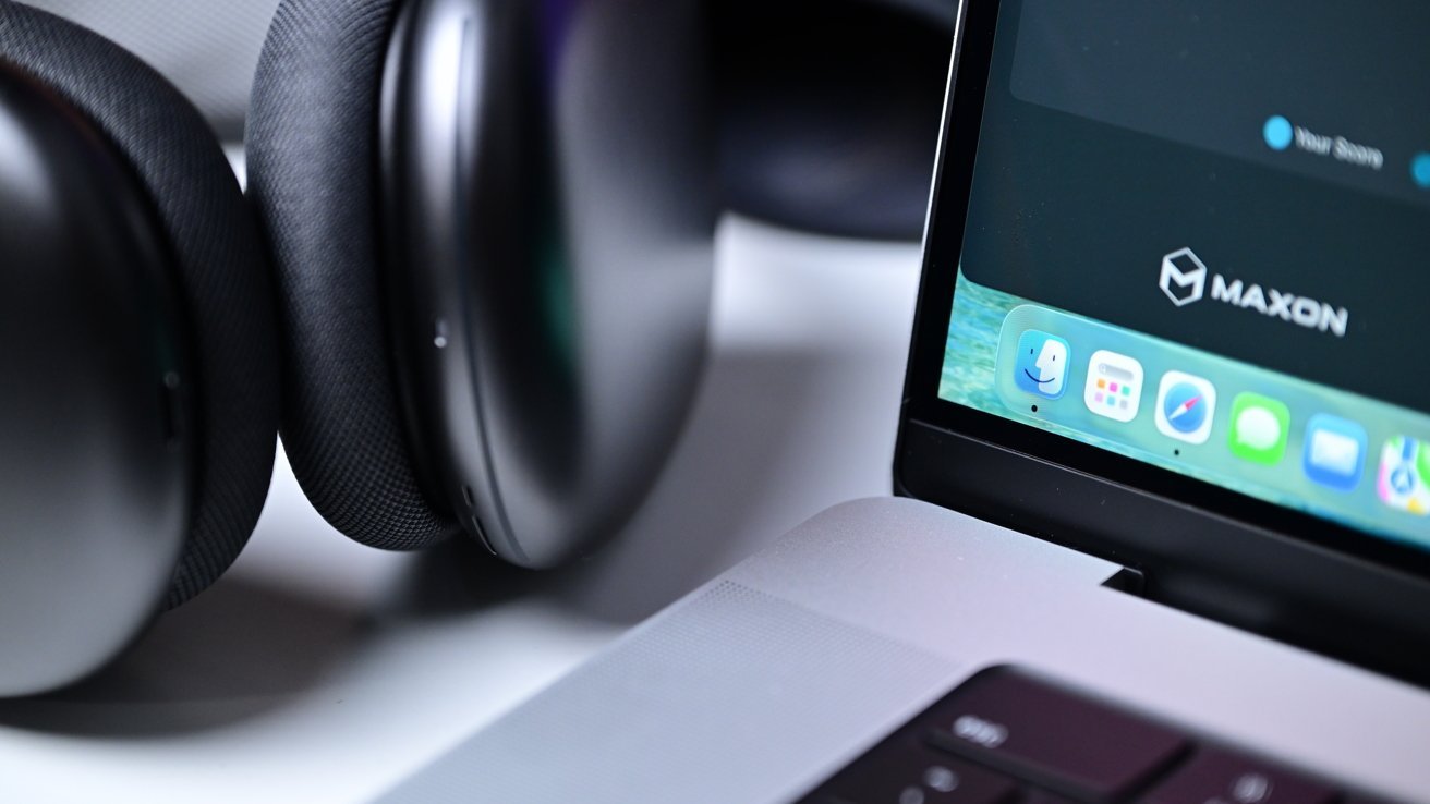 Close-up of AirPods Max resting beside an open MacBook Pro laptop, showing part of the keyboard, speaker grille, and colorful macOS dock on the screen