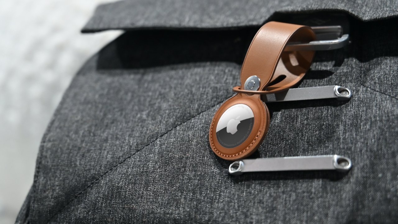 Apple AirTag in a tan leather keychain looped through a metal strap on a gray fabric bag or jacket, shown in a close-up view