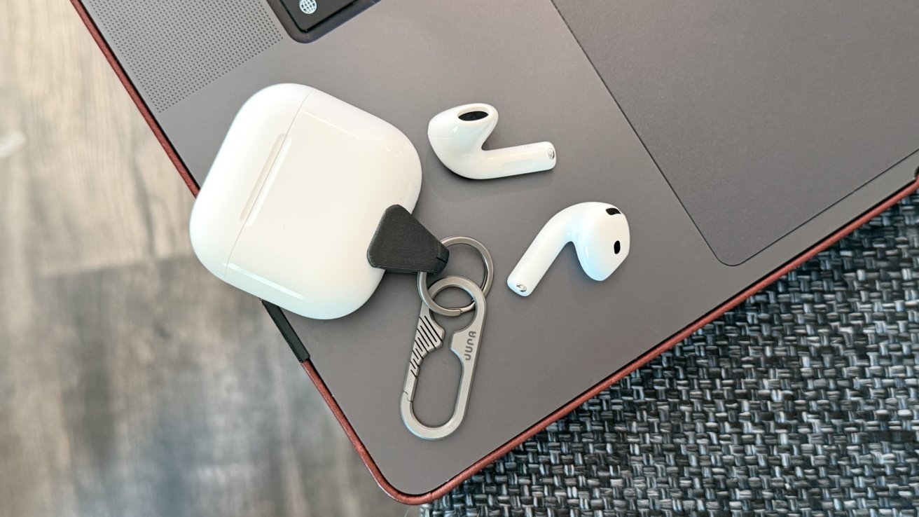 AirPods 4 earbuds, charging case, and a metal keychain with clip resting on the corner of a closed laptop on a textured dark woven surface