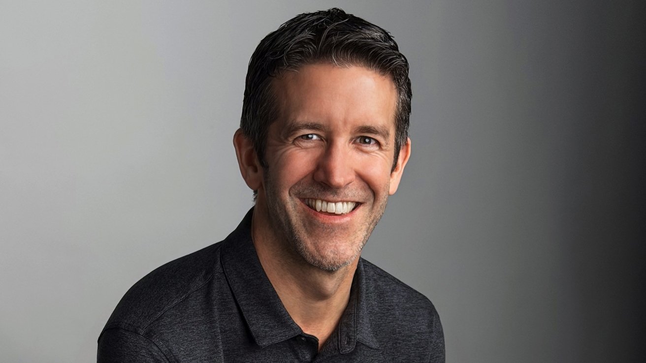 John Ternus, a smiling middle-aged man with short dark hair and light stubble wearing a dark polo shirt, posed against a simple gray background, looking directly at the camera
