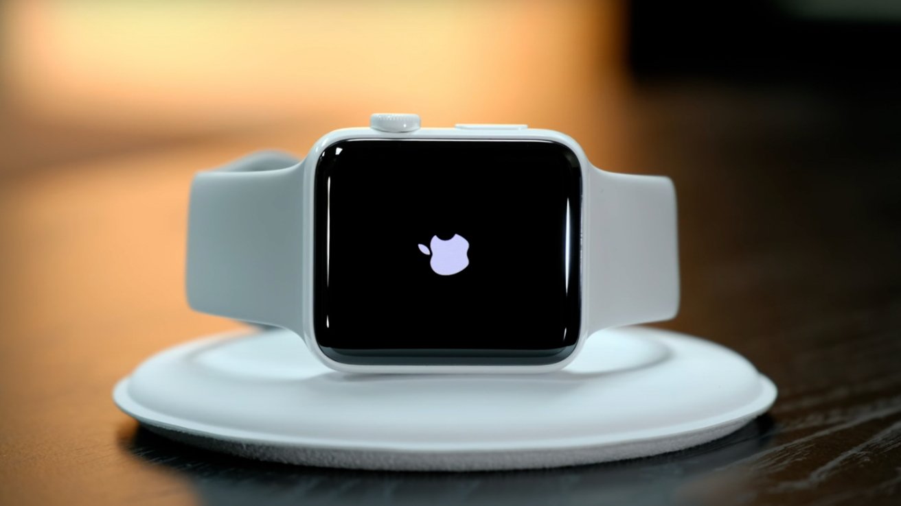 White Apple Watch with a black screen showing the glowing Apple logo, resting on a round white wireless charging pad on a wooden surface with a blurred warm background